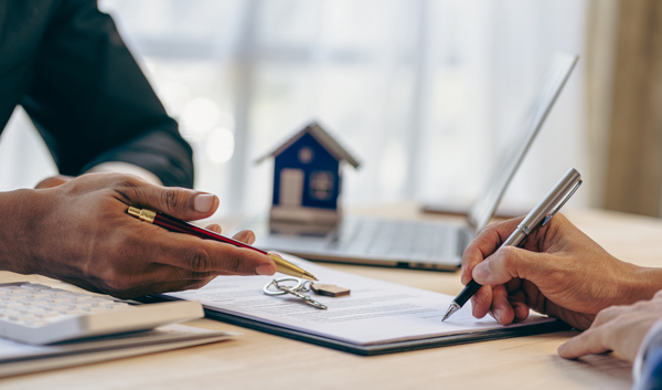 two hands signing a document with a pen and pencil on a table with a calculator and model house in the background document signing process real estate transaction