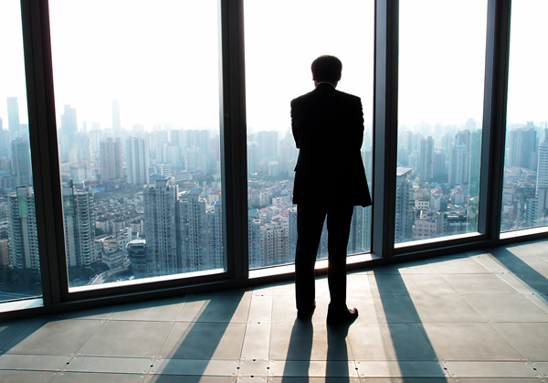 man in suit looking out window at city skyline with tall buildings and bright light reflecting on glass