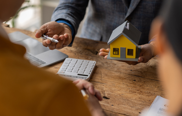 a person holding a small yellow house model with a calculator and laptop on the table representing property investment and real estate